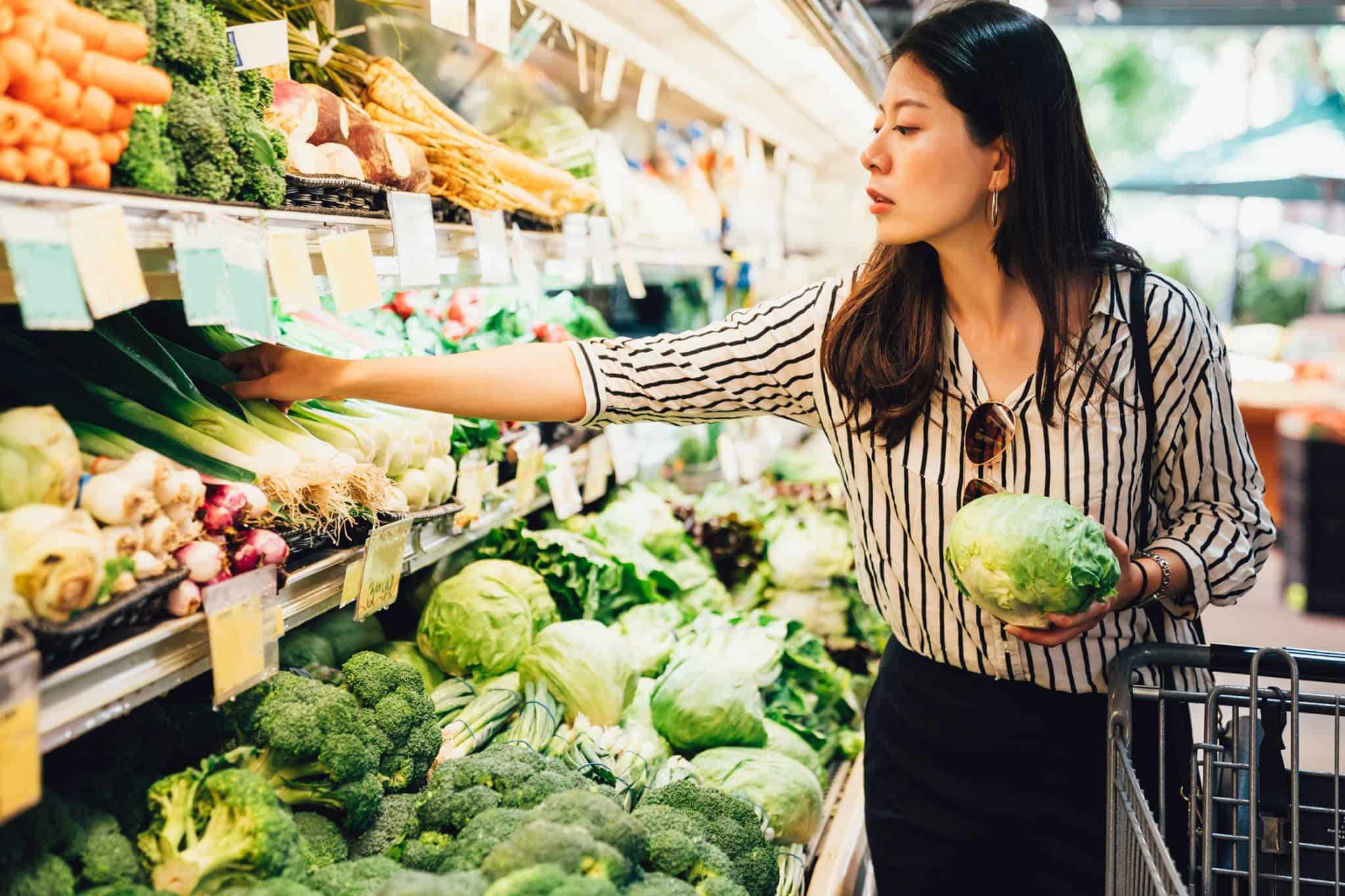A woman at the grocery store holding a head of lettuce while looking for other vegetable produce.