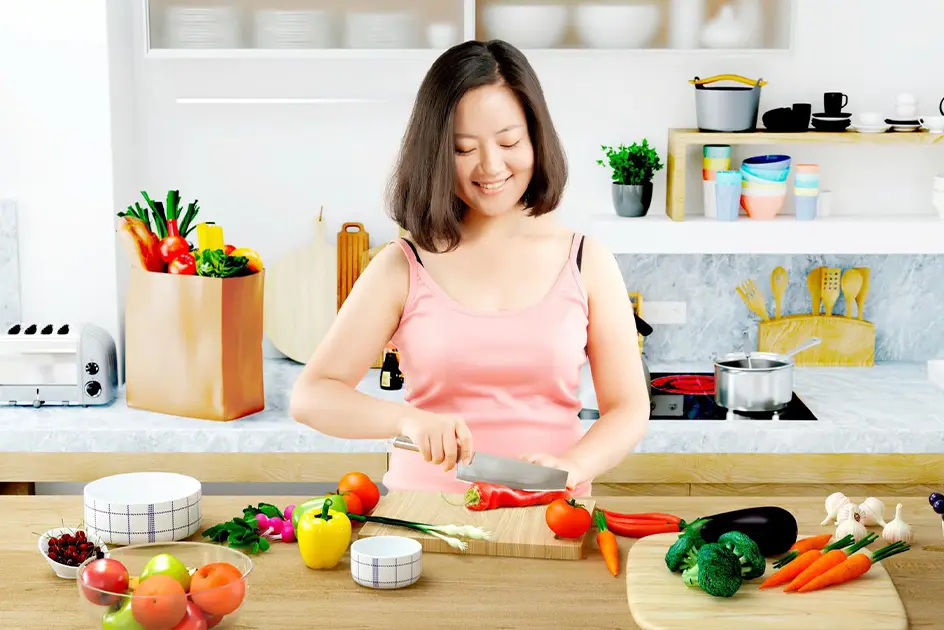 Woman in a kitchen chopping a pepper while being surrounded by other fruits and vegetables.