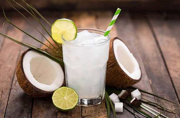 An iced drink with a green straw surrounded by coconuts and limes.