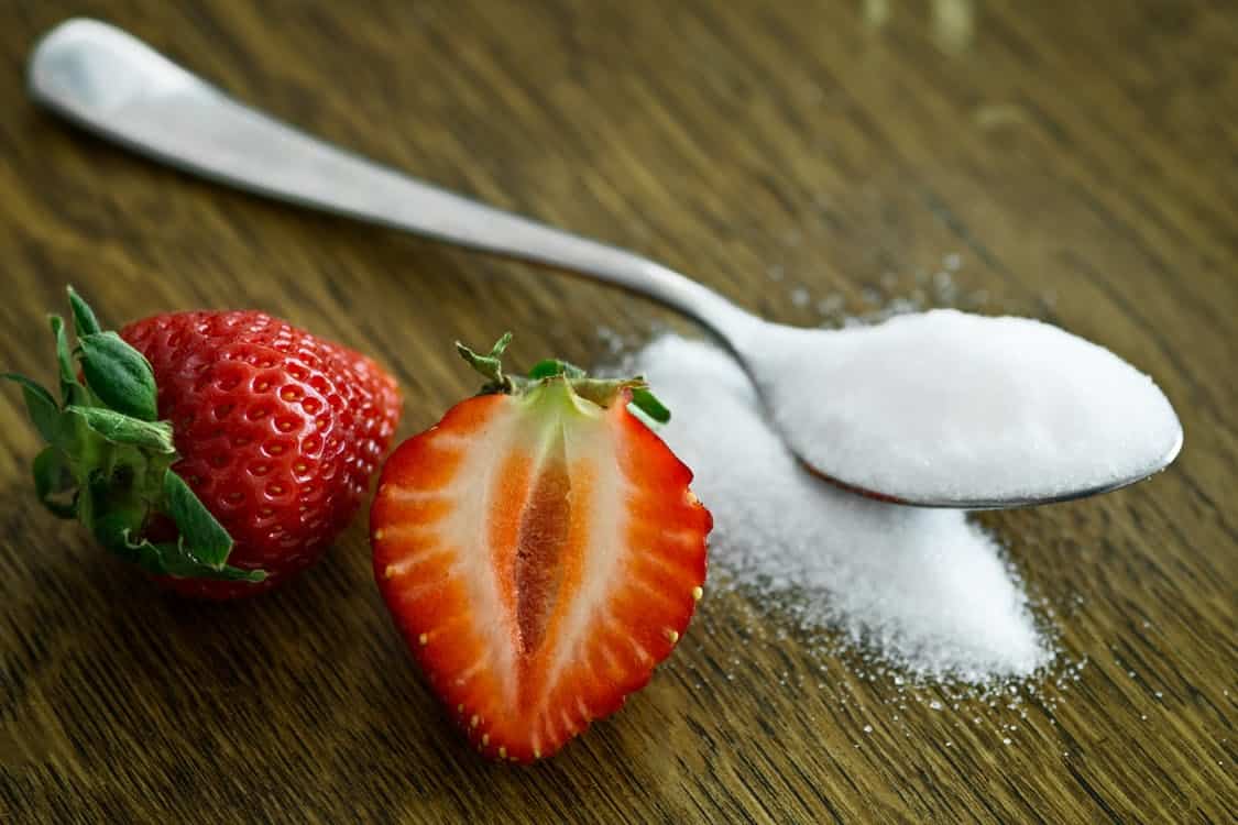 A pair of strawberries next to a spoonful of sugar.