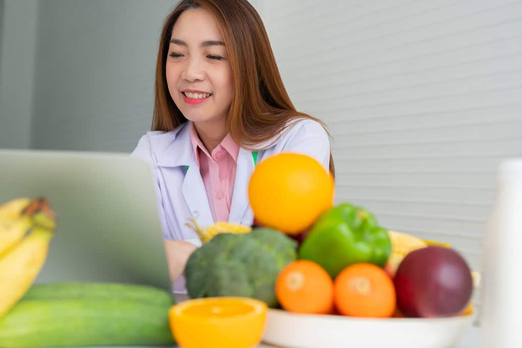Portrait of Asian smiling female nutritionist typing on laptop computer for balanced corrective diet plan for patient or weight loss advice. Concept of healthy lifestyle, healthy food, dieting